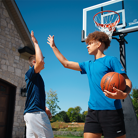 Two males models giving each other a high five in front of a Spalding basketball hoop. One of the models is holding a Spalding Zi/O Basketball. 